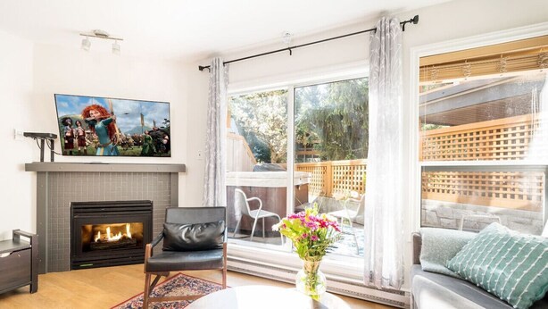 A living room with a fireplace, a sofa, a coffee table, and a large window with a view of the outdoors. - Host Large Groups w/Best Location+Private Hot Tubs (Whistler)