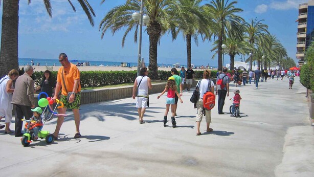 Eine belebte Promenade mit Palmen, ein Strand im Hintergrund und Spaziergänger.