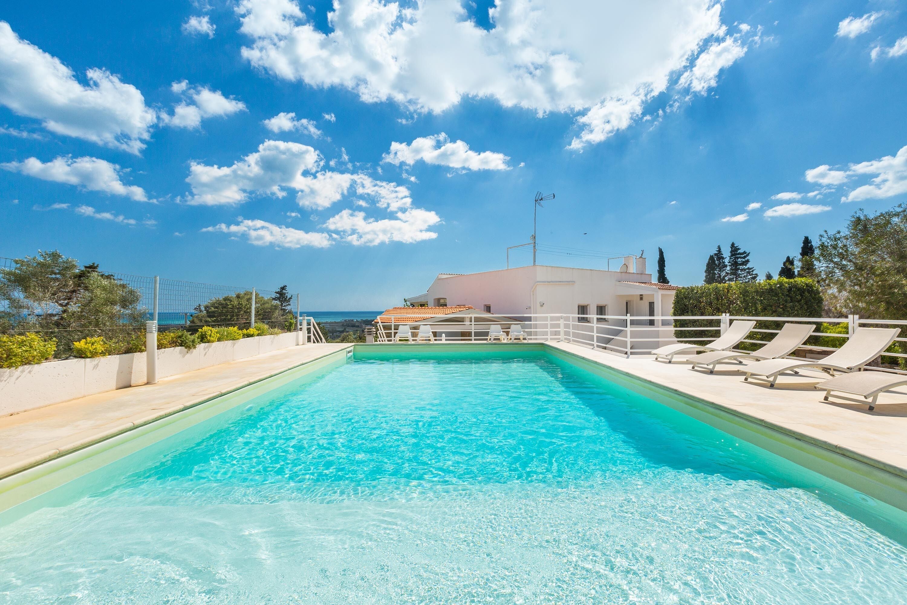 A rectangular swimming pool with clear blue water, surrounded by a white deck and lounge chairs. In the background, there is a white building with a red-tiled roof and a view of the sea.