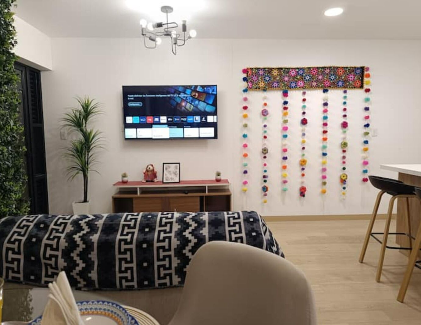 A modern living room with a flat-screen TV, a decorative wall hanging, a patterned sofa, and a small table with a plant.