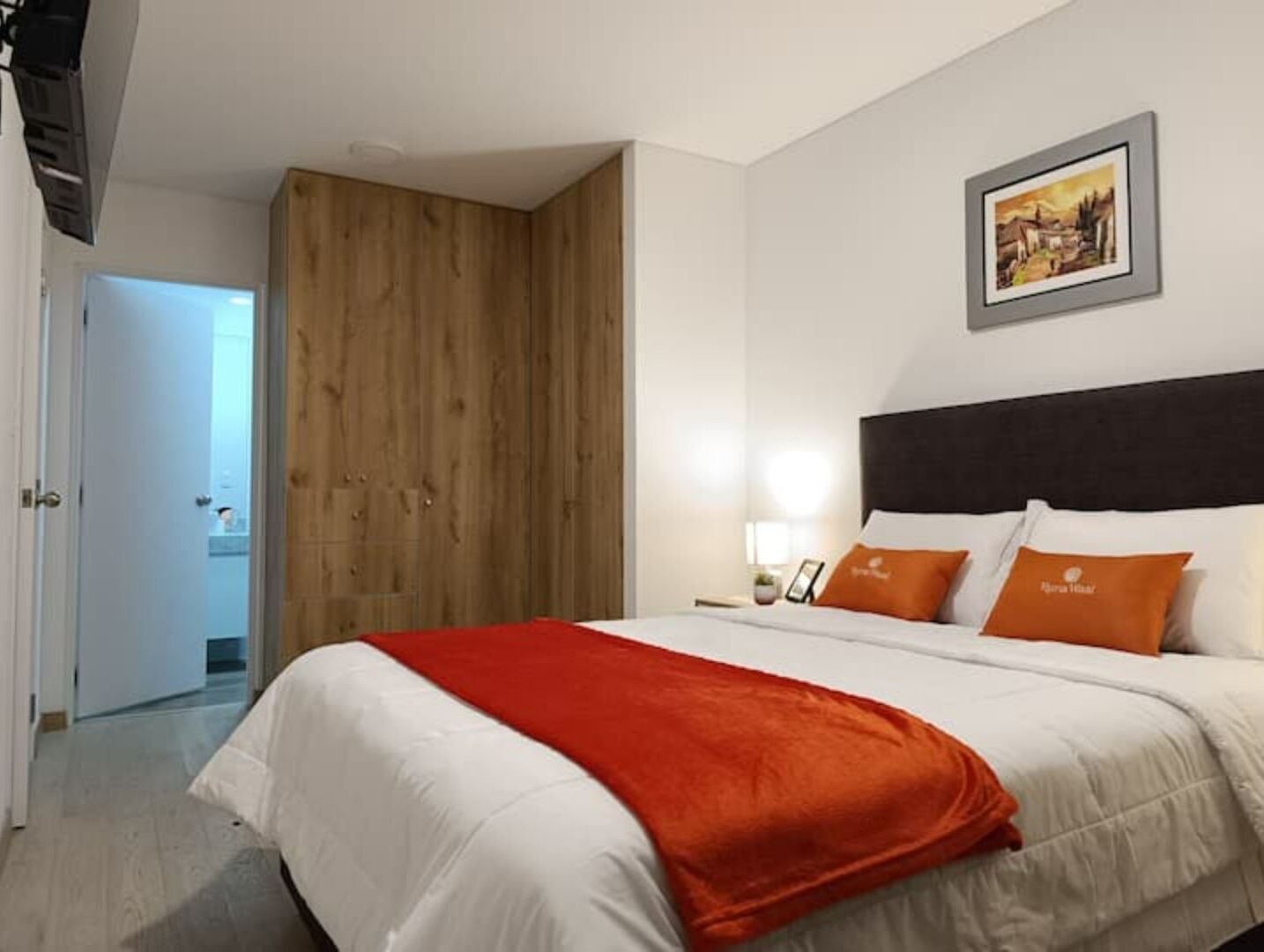 A hotel room with a bed, a wooden wardrobe, a nightstand with a lamp, and a framed picture on the wall.