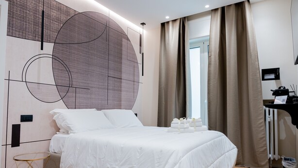 A modern hotel room with a large bed, a round wall decoration, a desk with a computer, and a window with curtains. - Calvani 23 Luxury House (Bari)