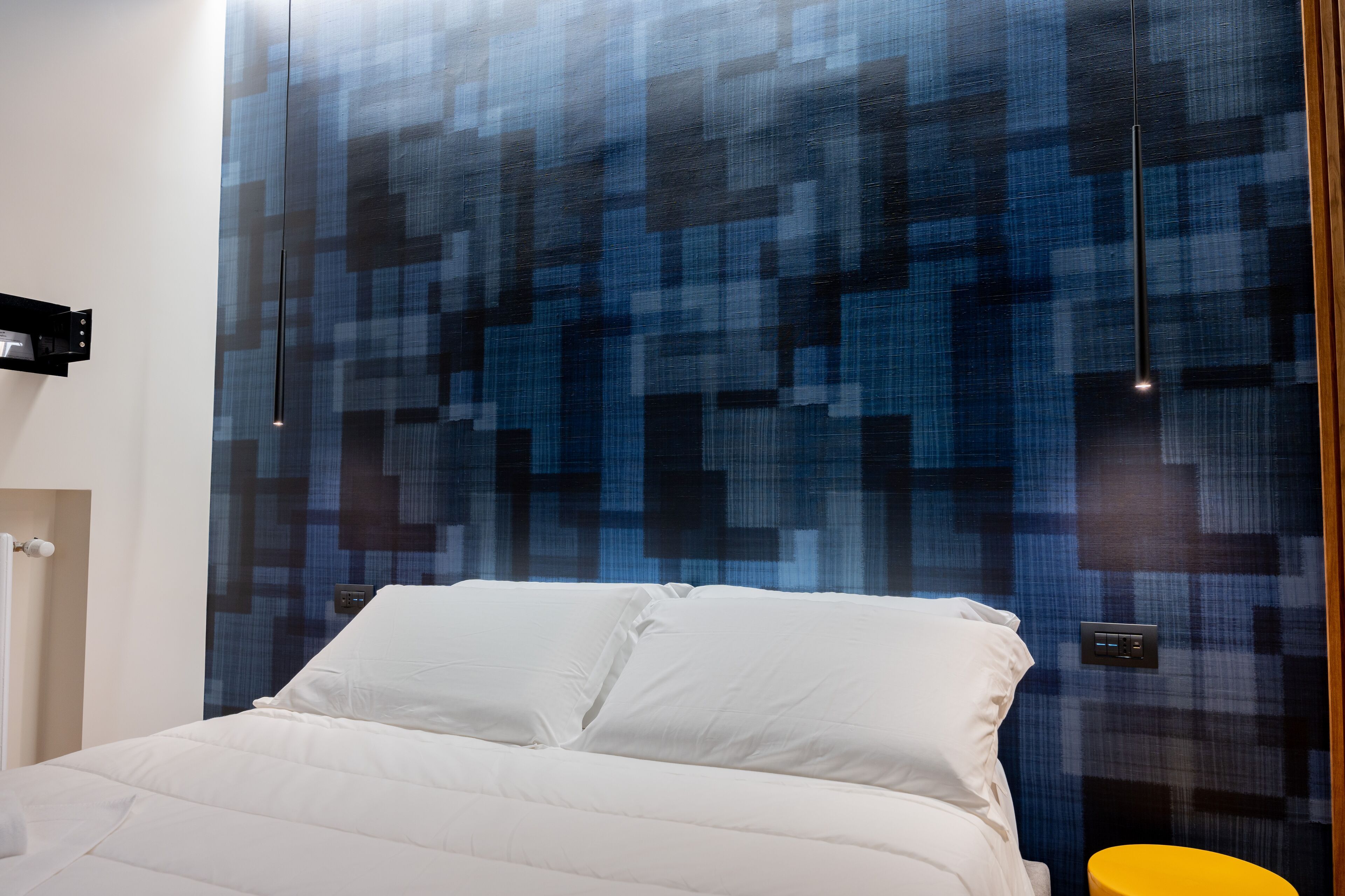 Una moderna camera d'albergo con un letto grande, una parete azzurra con motivo decorativo e un tavolino laterale giallo.