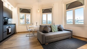 A modern apartment with a kitchenette, a grey sofa with patterned cushions, a dining table, and large windows offering a view of the city. - Matilde Modern apartment (Rome)