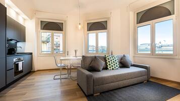Een modern appartement met een kitchenette, een grijze bank met gekleurde kussens, een eettafel en grote ramen met uitzicht op de stad.
