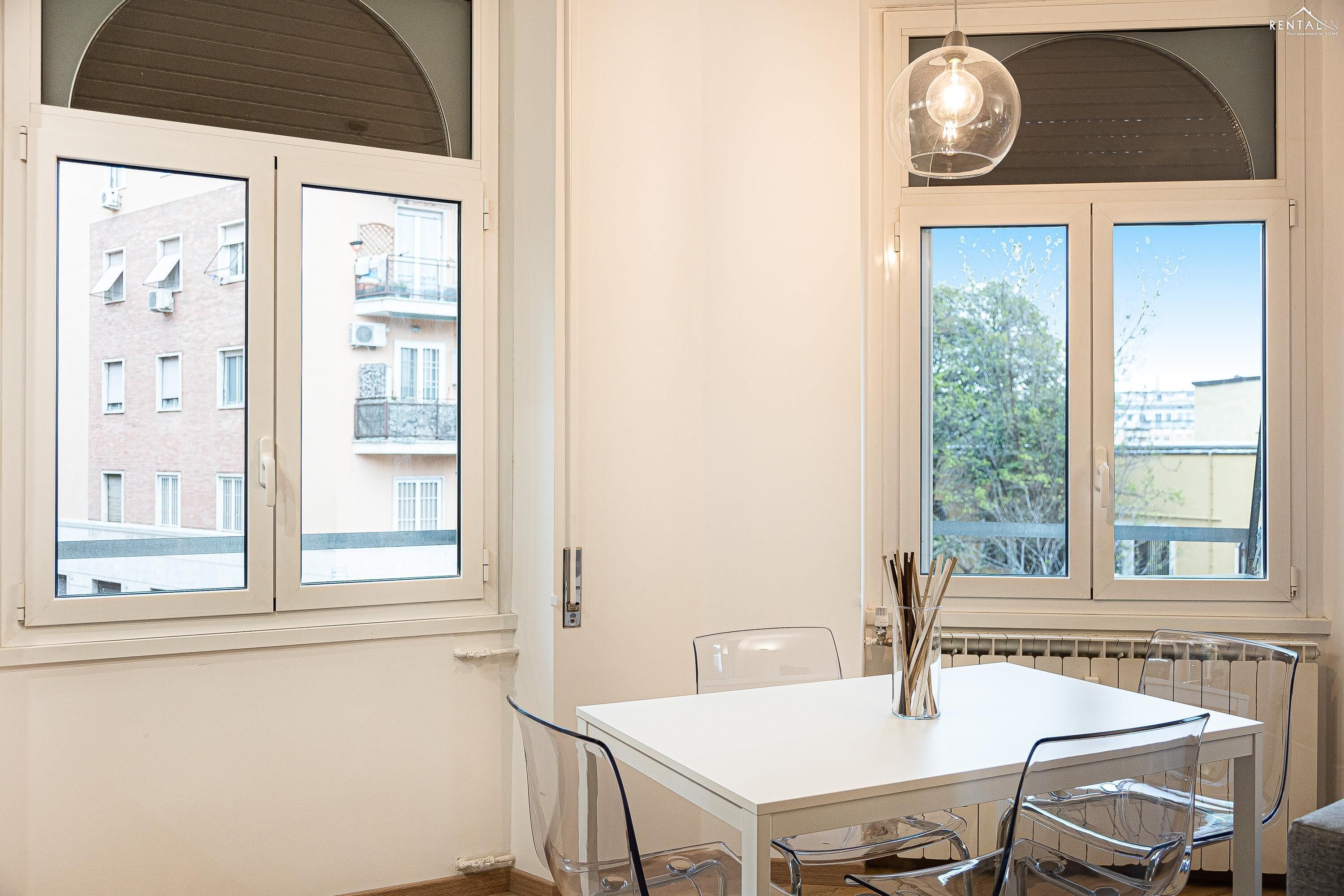 Een moderne eetruimte met een witte tafel en stoelen, grote ramen met uitzicht op gebouwen en bomen, en een hanglamp.