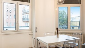 A modern dining area with a white table and chairs, large windows with views of buildings and trees, and a hanging light fixture. - Matilde Modern apartment (Rome)