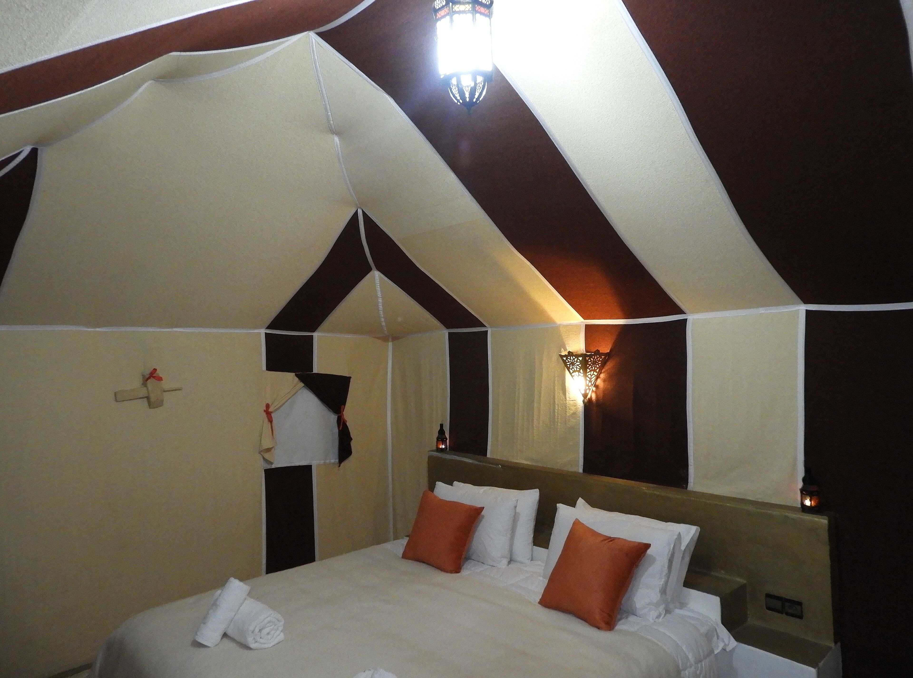 A bedroom with a high ceiling, a bed with white bedding and orange pillows, a hanging light fixture, and a mirror on the wall.