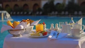 A breakfast setup by a pool with a table set for two, including a bowl of fruit, a plate of eggs, and a pitcher of orange juice. - Luxury  Saf Saf Camp (Taouz)