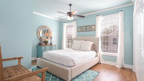 Une chambre à coucher comprenant un lit, une commode, un miroir, un ventilateur de plafond et une fenêtre avec des rideaux.