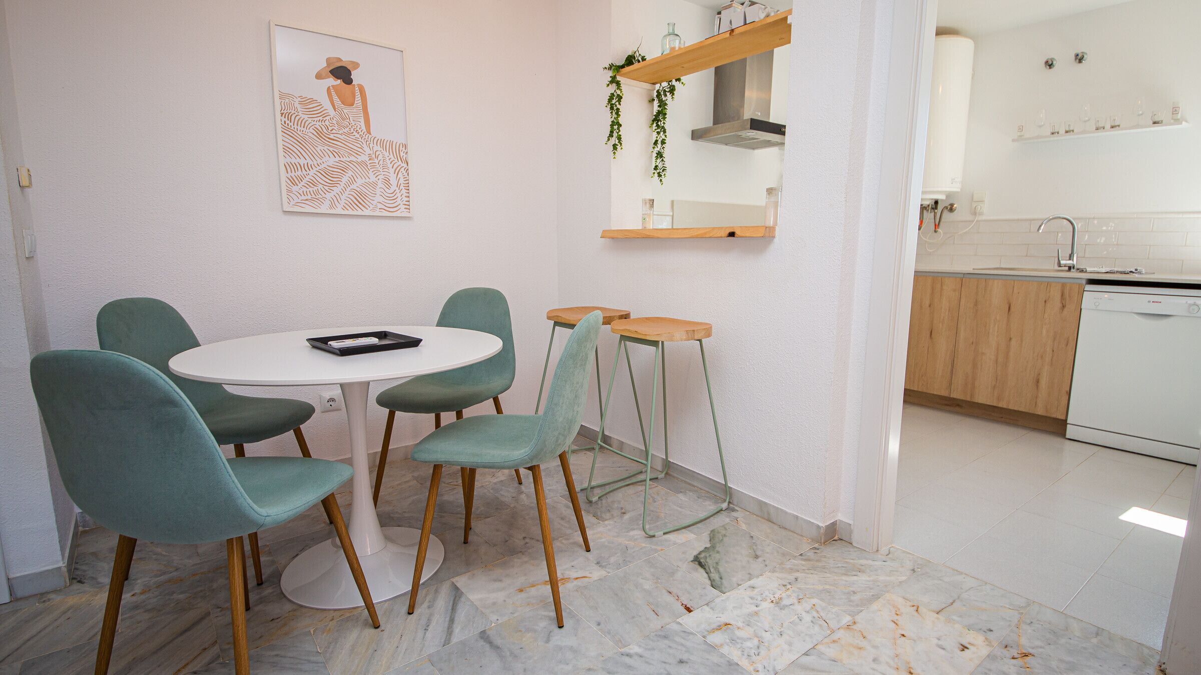 A small dining area with a round table and four chairs, a kitchen with white cabinets and a dishwasher, and a wall-mounted shelf with plants.