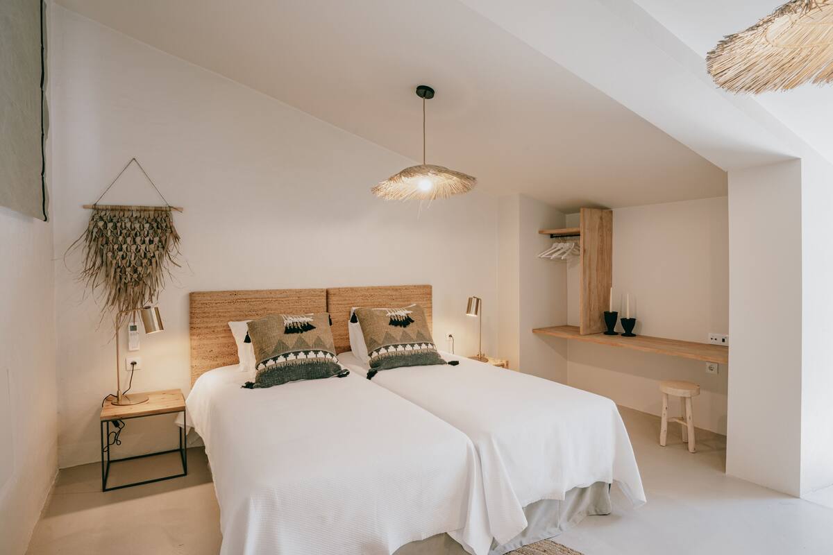 A modern bedroom with two beds, a hanging light fixture, a bedside table, and a small stool.