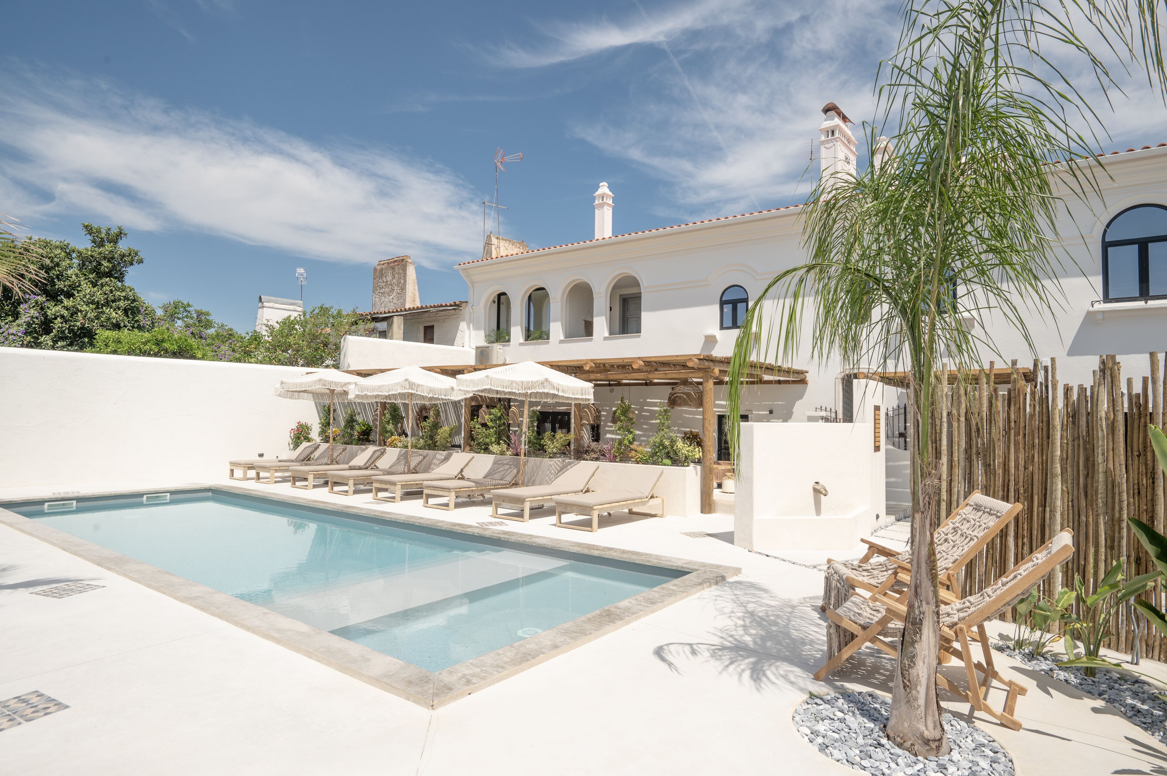 Una piscina con sillas de descanso y sombrillas, rodeada por un edificio blanco con ventanas de arco y una chimenea.