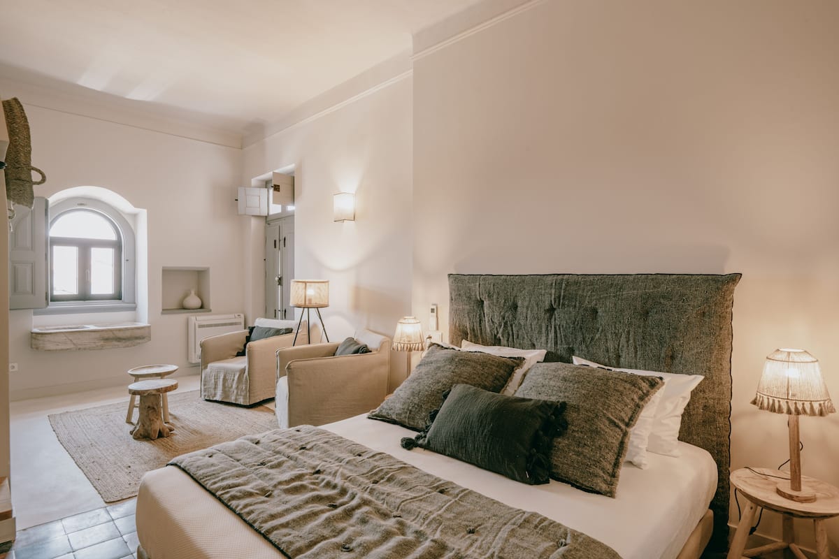 A bedroom with a large bed, two bedside tables with lamps, a chair, a small table, and a window with a view.