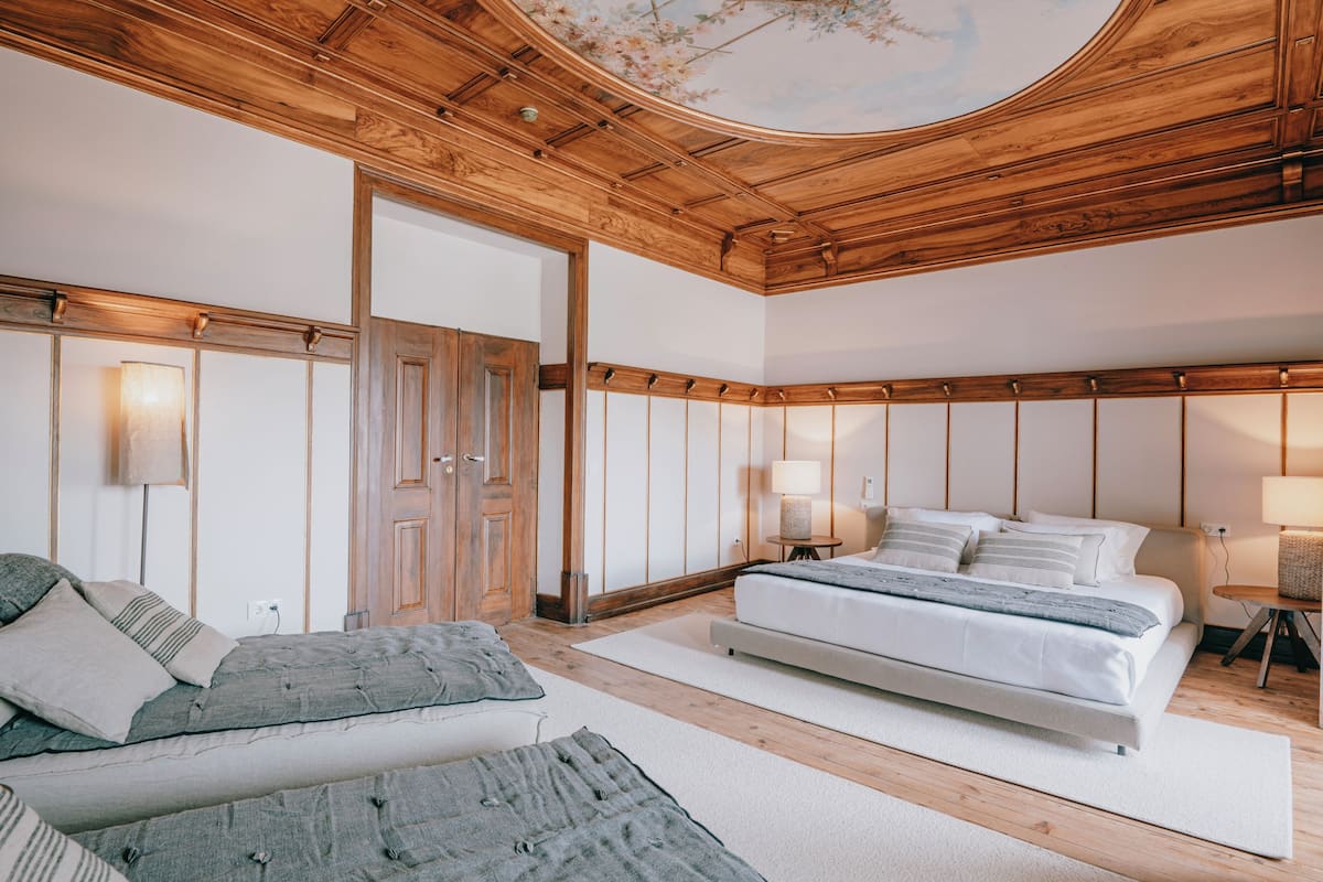 A bedroom with a wooden ceiling, a large bed, two bedside tables with lamps, and a door.