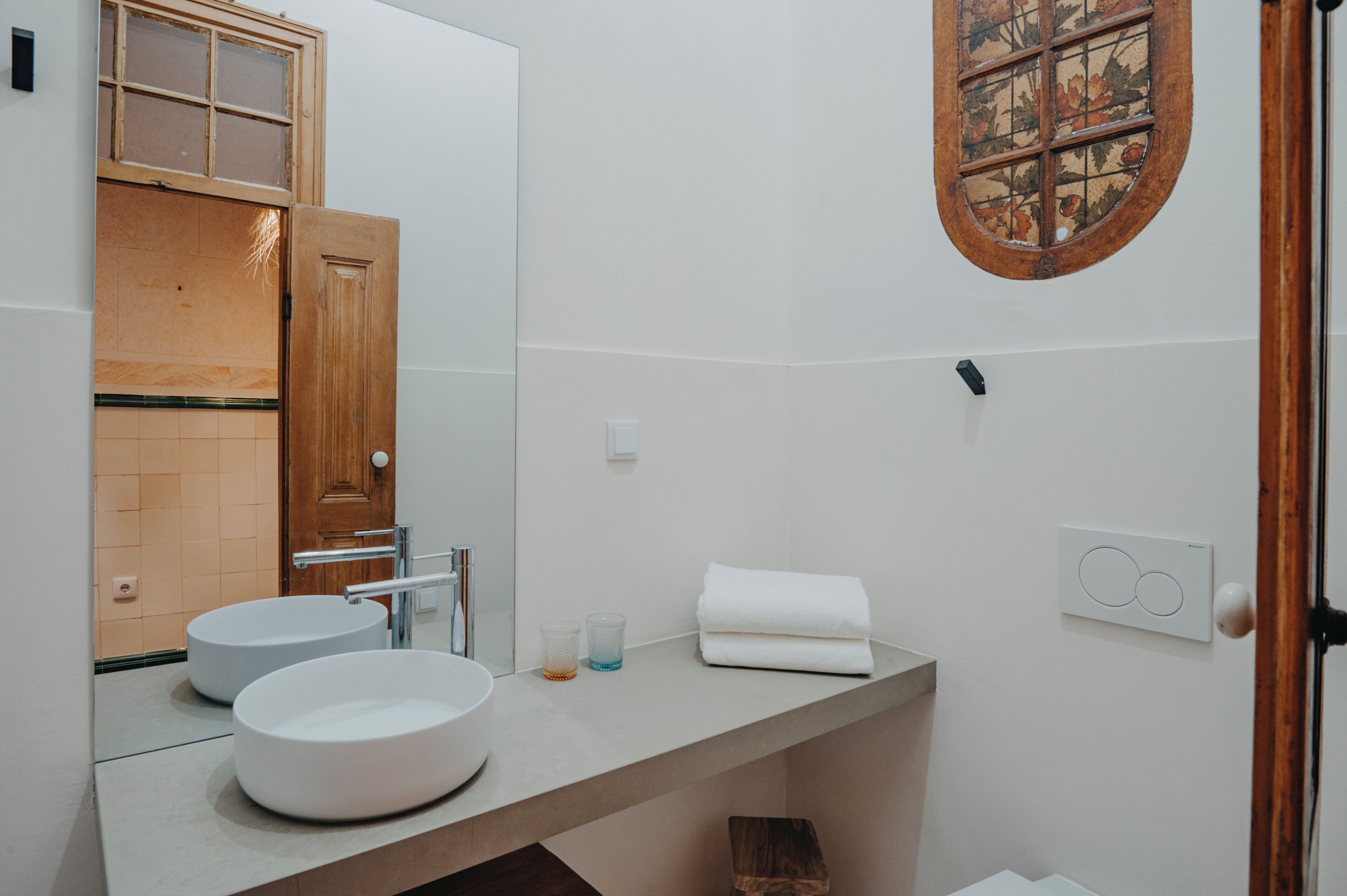 Un baño con dos lavamanos redondos, una puerta de madera, un espejo y un ventanal decorativo.