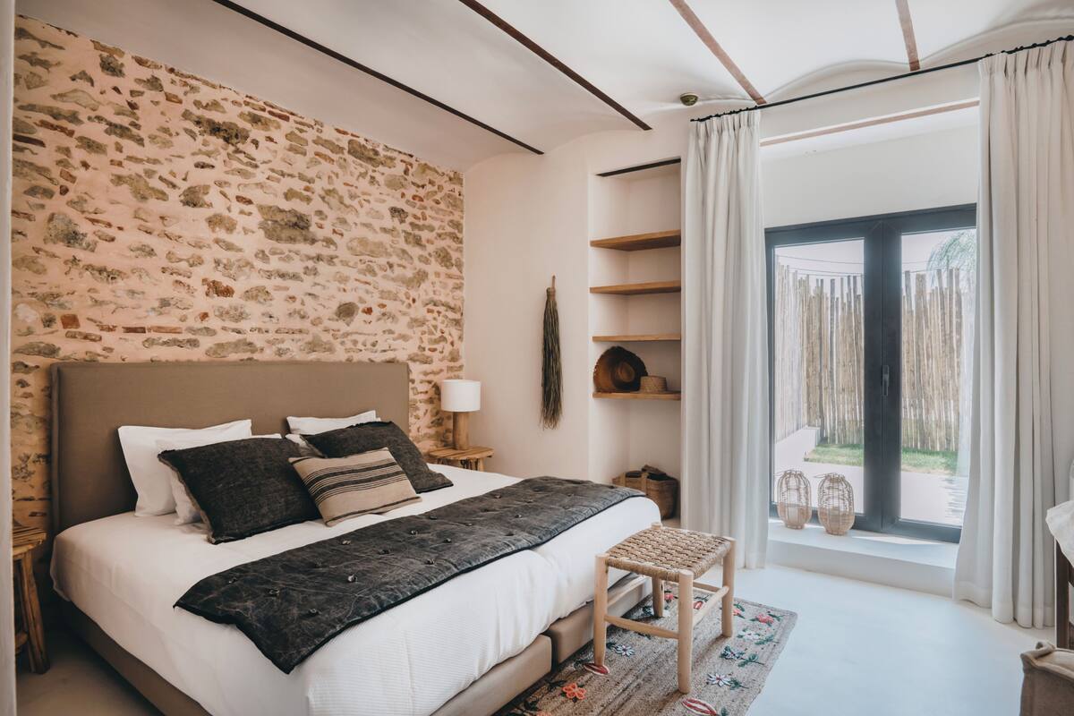 A bedroom with a stone wall, a bed with a dark headboard, a bedside table with a lamp, and a window with curtains.