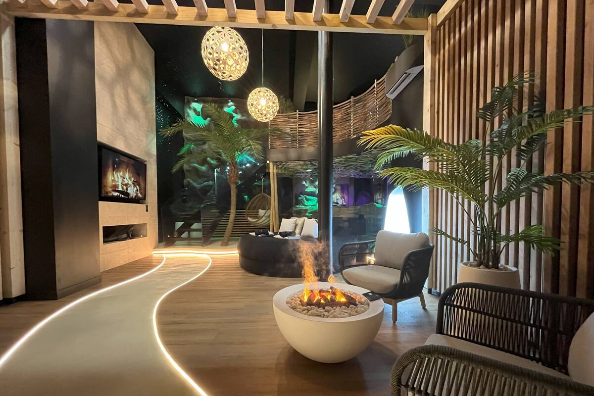 A modern lounge area with a curved wooden floor, a central fire feature, seating, and hanging spherical lights.