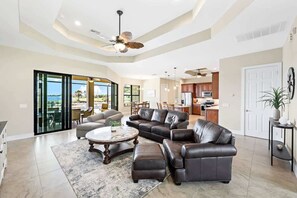 A spacious living room with a large sofa, ottoman, and coffee table. There is a ceiling fan, a kitchen area, and a sliding glass door leading to a patio.