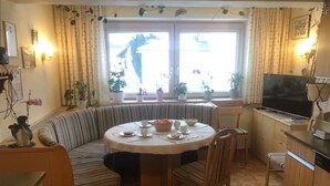 Un comedor acogedor con una mesa redonda, sillas y un banco empotrado. Hay un televisor a la derecha y plantas en macetas en el alféizar de la ventana.