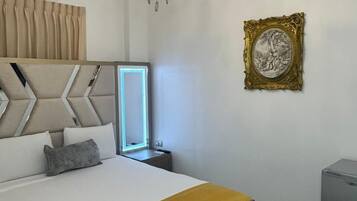 A bedroom with a bed, a bedside table, a nightstand, a mirror, a chandelier, a picture frame, and a small cabinet.
