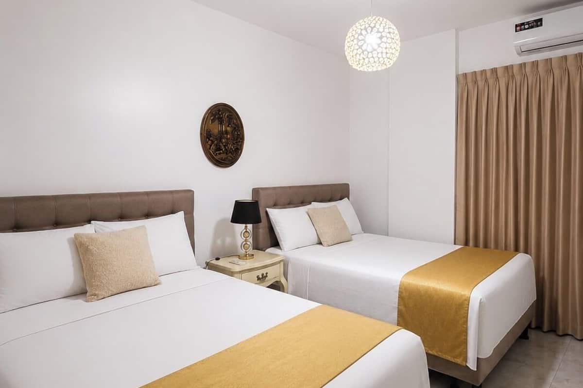 A hotel room with two beds, a nightstand, a wall clock, a ceiling light fixture, and an air conditioning unit.