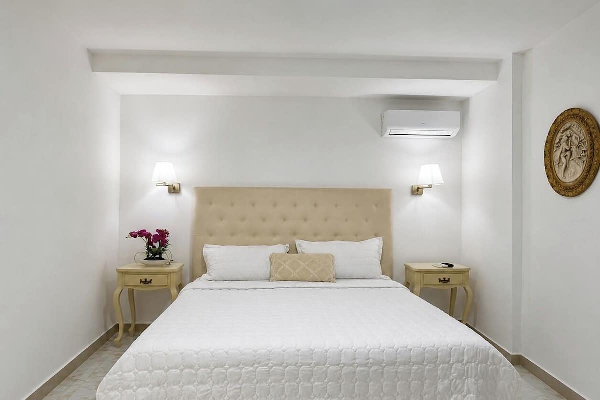 A bedroom with a large bed, two bedside tables, a wall-mounted air conditioner, and a decorative plate on the wall.