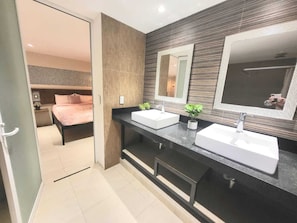 A modern bathroom with two sinks, a large mirror, and a bed visible through an open door.