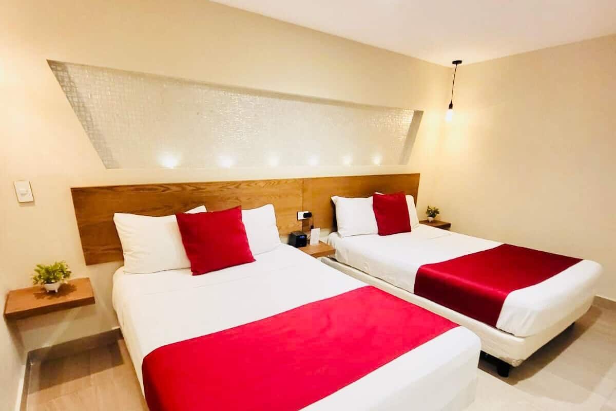A hotel room with two beds, each with white bedding and red accents, a wooden headboard, a small bedside table with a plant, and a large window with a view.