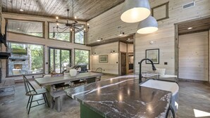 A spacious kitchen with a large island, wooden ceiling, and large windows offering views of the outdoors.