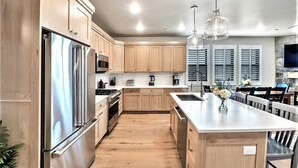 A modern kitchen with wooden cabinets, stainless steel appliances, a central island, pendant lights, and a stone wall with a TV.