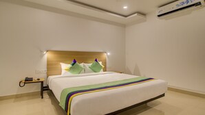 A hotel room with a wooden headboard, a bed with a white bedspread and colorful pillows, a bedside table with a phone and remote control, and an air conditioning unit on the wall. - Treebo De Alphabet Kondapur (Hyderabad)