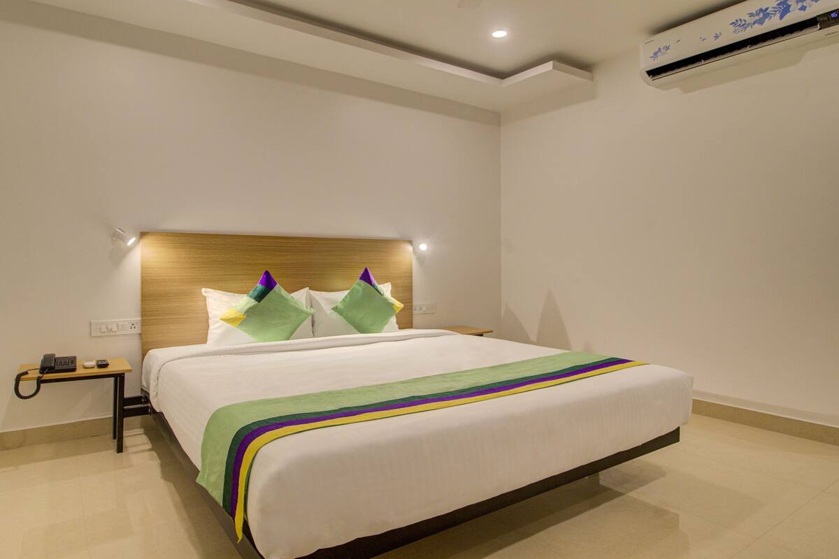A hotel room with a wooden headboard, a bed with a white bedspread and colorful pillows, a bedside table with a phone and remote control, and an air conditioning unit on the wall.