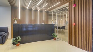 A reception area with a black reception desk, two potted plants, a grey sofa, and a wooden wall with vertical slats. - Treebo De Alphabet Kondapur (Hyderabad)