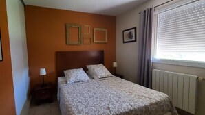 Una habitación de hotel con cama, dos mesitas de noche, ventana con persianas e imágenes enmarcadas en la pared.