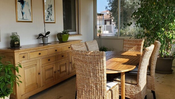 Un comedor con mesa de madera y sillas de mimbre, un mueble de madera con plantas y un ventanal con vista a una casa y árboles.