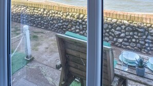 A view through a window of a patio area with a stone wall, a wooden bench, and a table with a potted plant and glassware. - 1 bedroom accommodation in Sheringham (Sheringham)