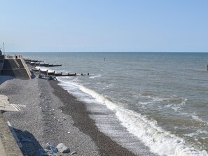 A pebbled beach with a staircase leading down to the water, a wooden jetty extending into the sea, and a clear sky.