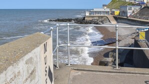 A coastal promenade with a metal railing, a rocky shoreline, and buildings in the background. - 1 bedroom accommodation in Sheringham (Sheringham)