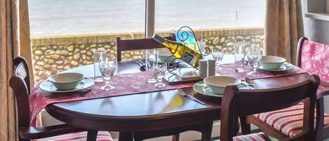 A dining table set for two with a view of the sea, a bottle of wine, and a patterned tablecloth.