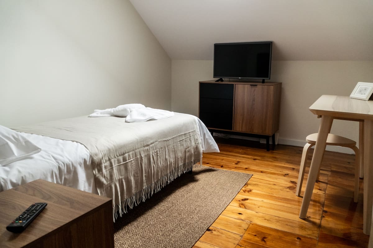 Ein Schlafzimmer mit einem Bett, einem Fernseher auf einer Holzkommode, einem Schreibtisch mit Stuhl sowie einer Fernbedienung auf einem Nachttisch aus Holz.