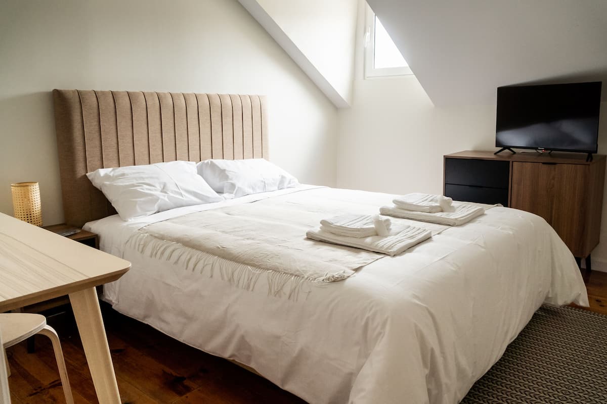 Ein ordentlich bezogenes Bett mit weißen Leinen und Handtüchern, ein Nachttisch aus Holz, ein Fernseher auf einer Holzkommode und ein kleiner Tisch mit einer Lampe.