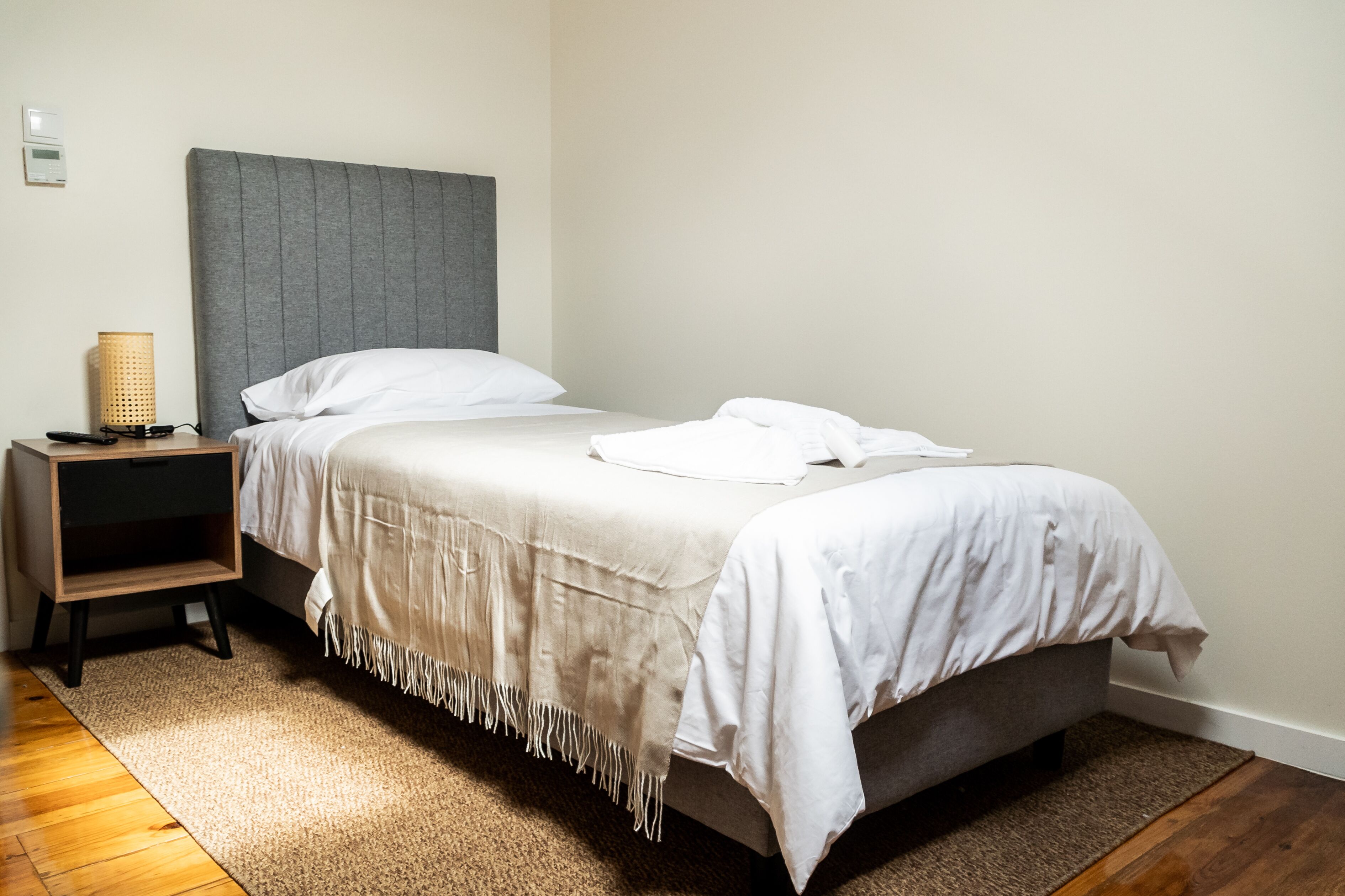 Uma cama bem arrumada com cabeceira cinza, uma mesa de cabeceira com abajur e piso de madeira.