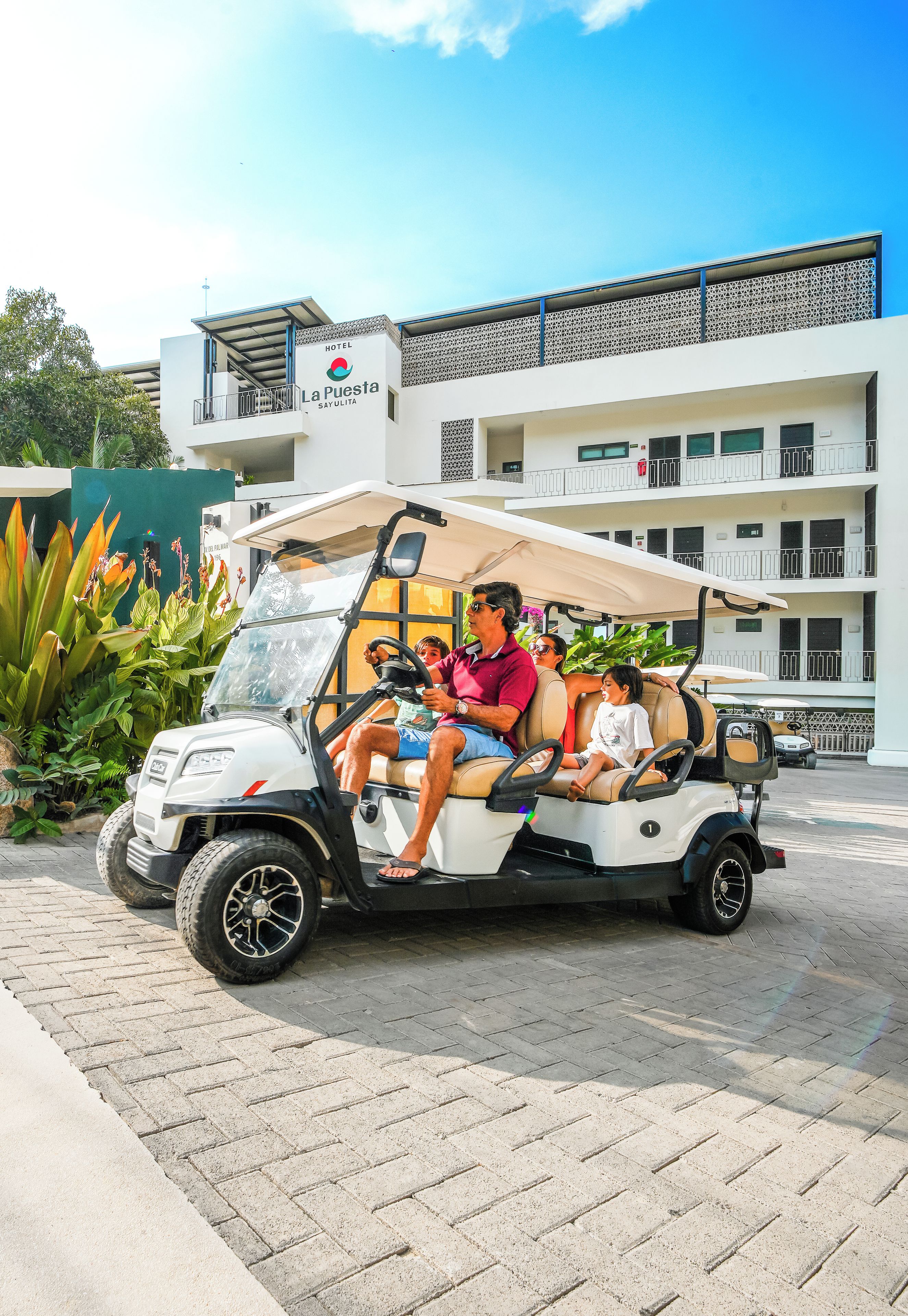 Un carrito de golf con pasajeros, estacionado frente a un hotel con un letrero que dice 'La Puesta Sayulita'.