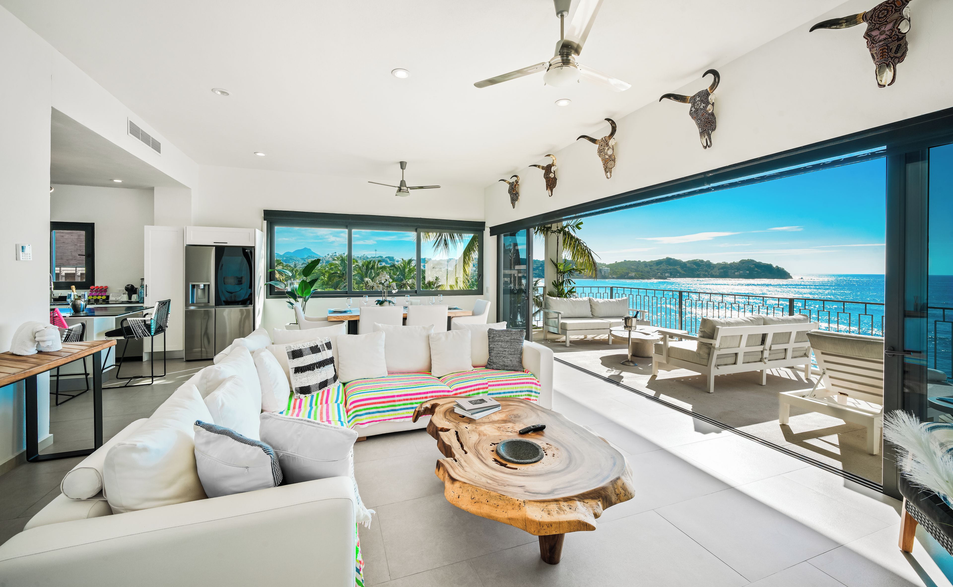 Una sala de estar moderna con un ventanal que ofrece vistas panorámicas al océano, un comedor con una cocina al fondo y un balcón con mobiliario exterior.