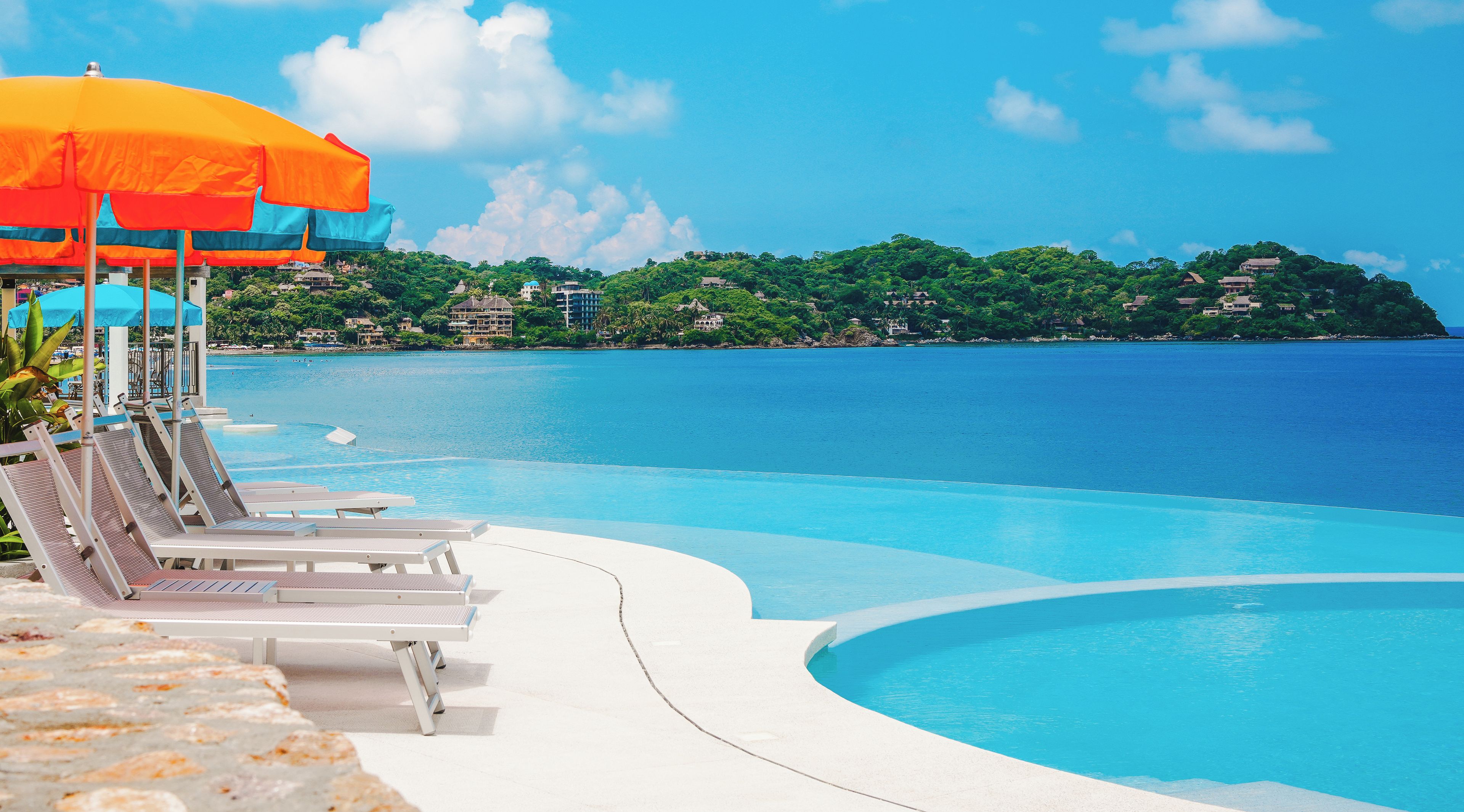 Piscina azul límpida com espreguiçadeiras e um guarda-sol, emoldurada por paisagem tropical e vista para o mar.