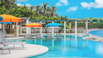 Un área de piscina con sillas de descanso y sombrillas, rodeada de palmeras y vista a la playa.