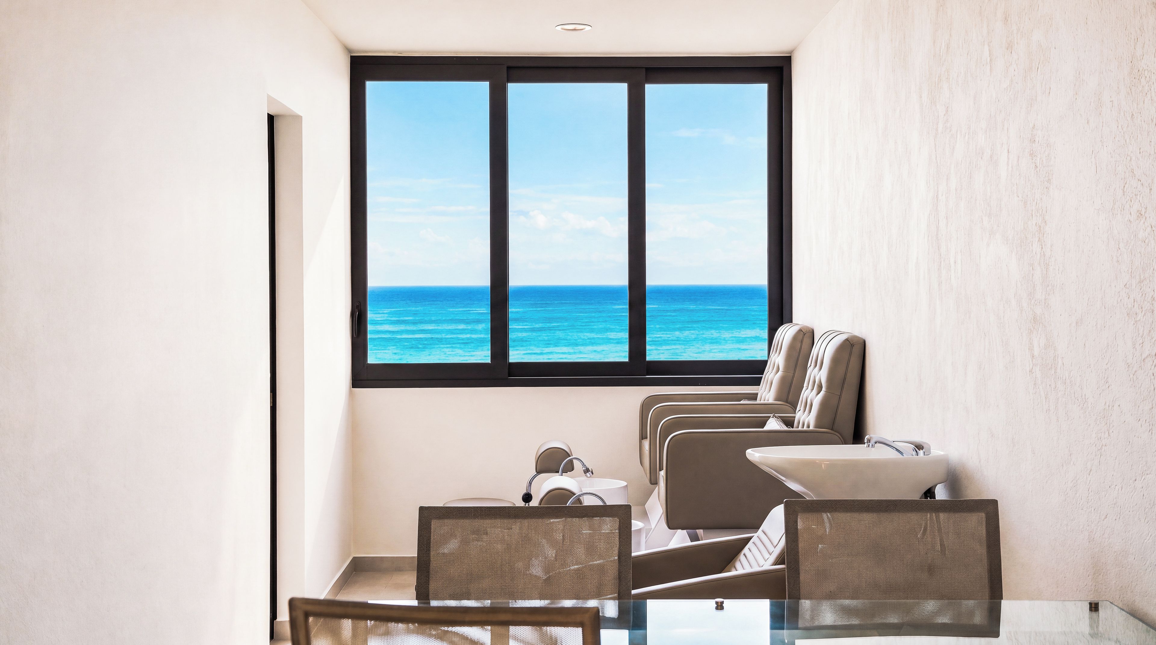 Una habitación con un amplio ventanal con vistas al océano, una mesa de vidrio y dos sillones acolchados.