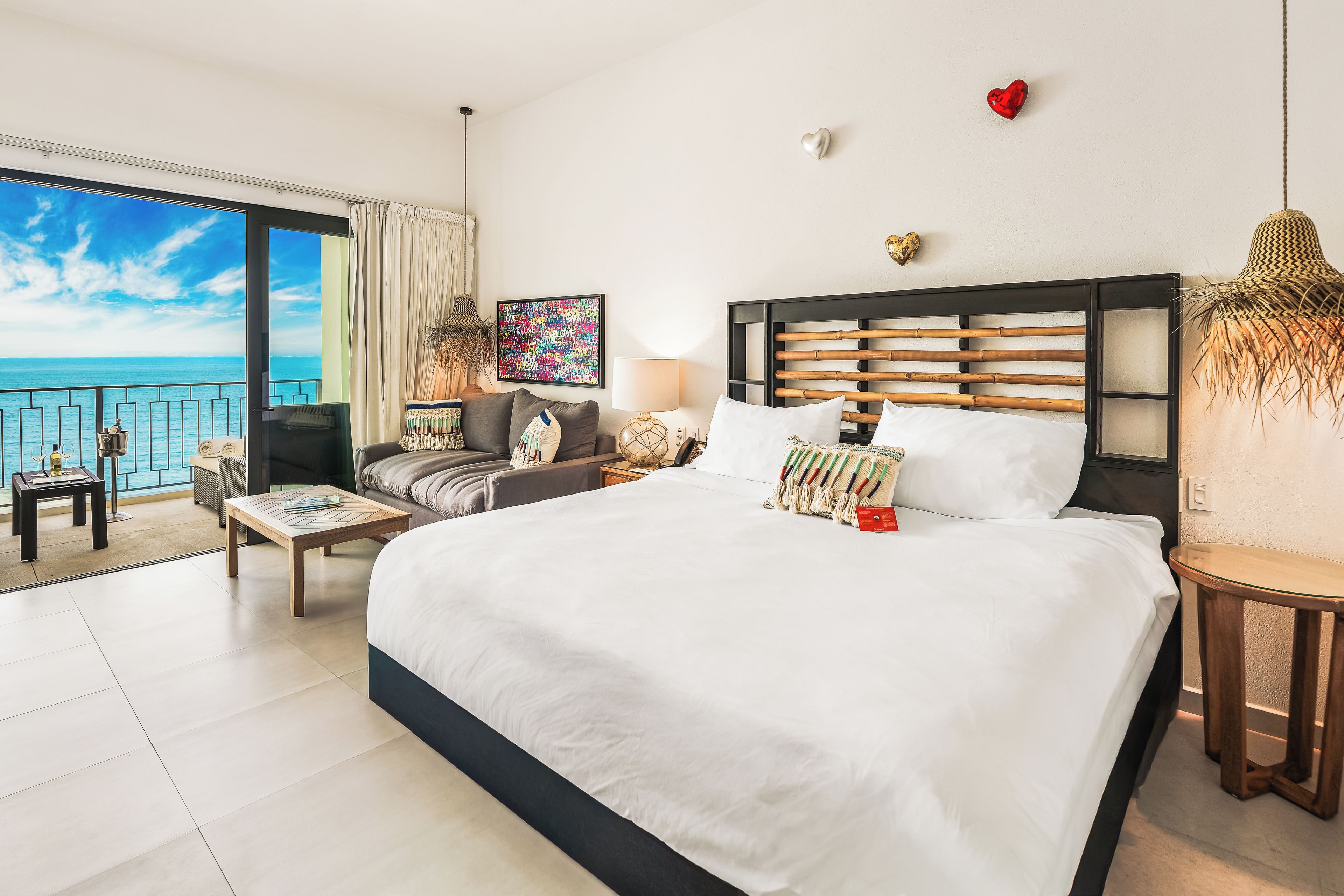 A hotel room with a large bed, a sofa, a small table, a chair, a lamp, and a view of the sea.