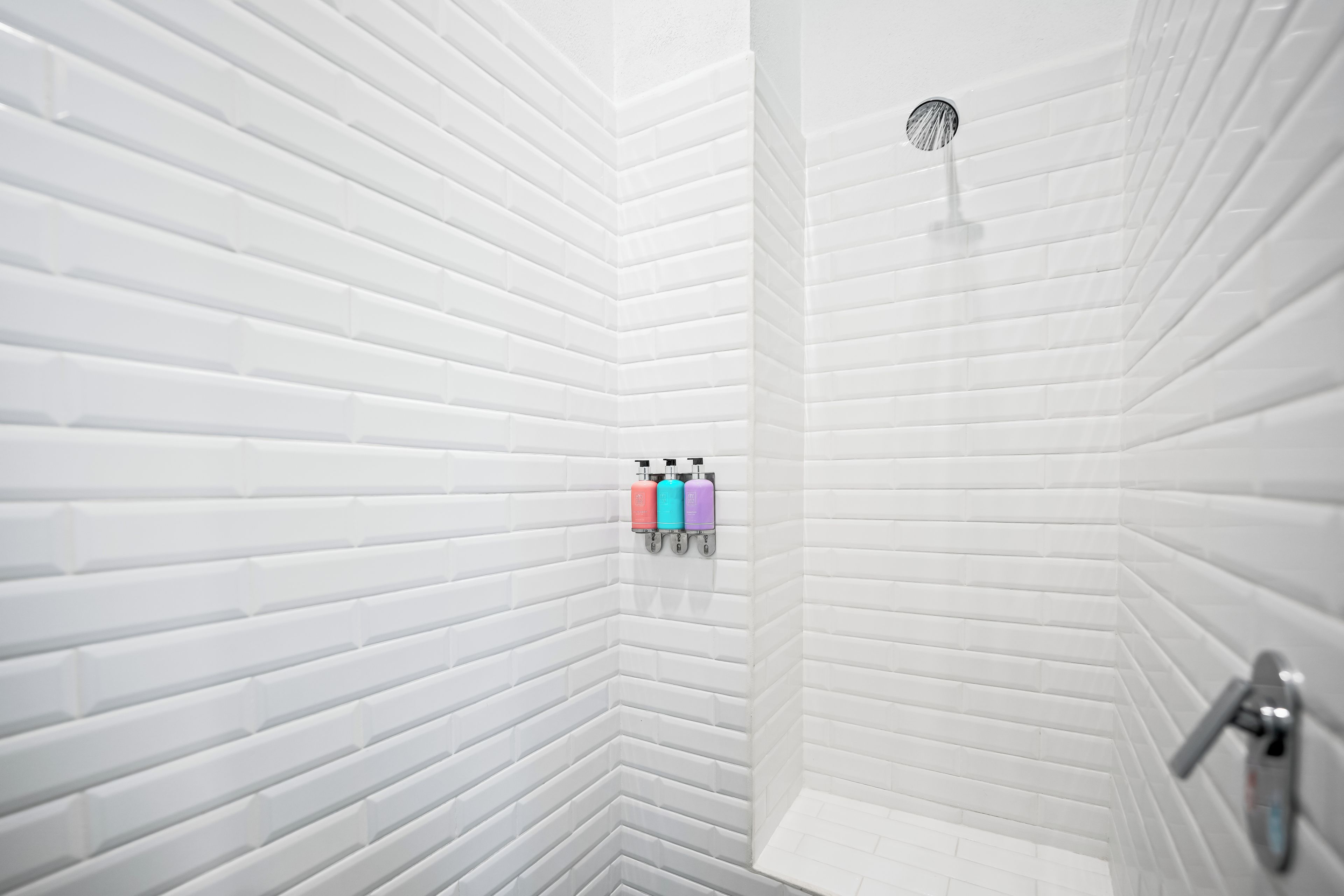 A corner of a bathroom with white tiled walls and a shower. There are two bottles of shampoo and conditioner on a wall-mounted dispenser.